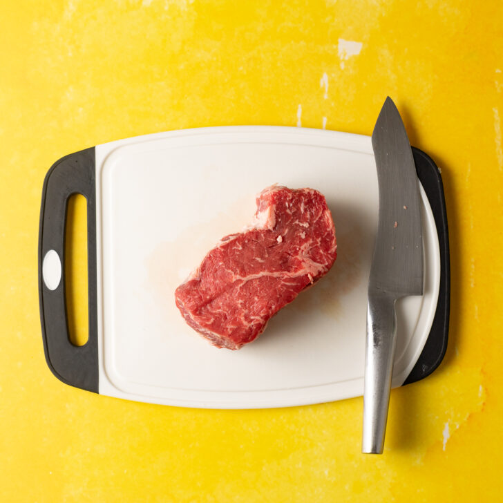 Ribeye steak on white cutting board with knife.