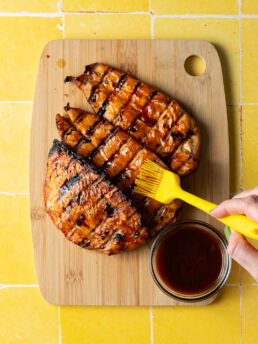 Marinated barbecue chicken on a cutting board being brushed with more barbecue sauce.