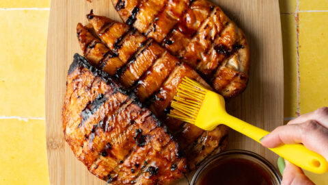 Marinated barbecue chicken on a cutting board being brushed with more barbecue sauce.