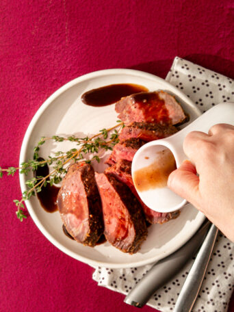 A hand pours rich bordelaise sauce over sliced roast beef arranged on a white plate with sprigs of fresh thyme, a polka-dot napkin, and utensils, all set on a red surface.