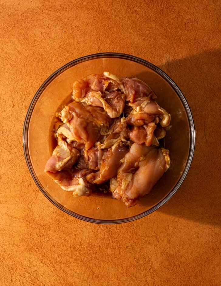 A glass bowl filled with raw, marinated chicken pieces sits on an orange-brown textured surface.