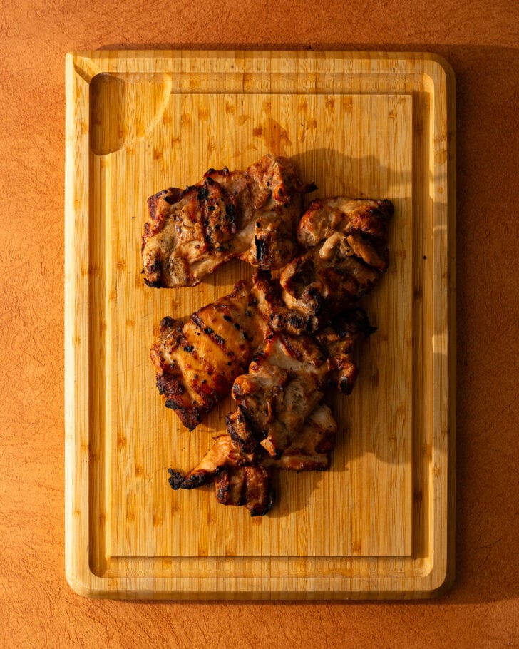 Grilled pieces of marinated chicken are placed on a wooden cutting board, with visible grill marks and a warm, sunlit background.