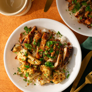 A white bowl with grilled chicken, roasted cauliflower, and white rice, topped with chopped herbs, sits on an orange tablecloth next to a cup and gold utensils.