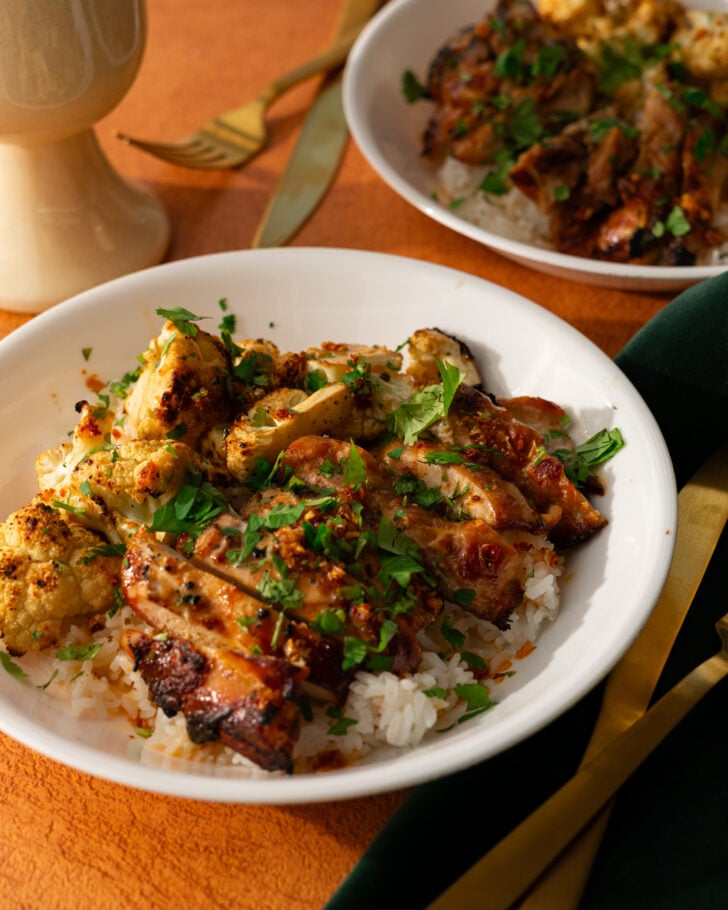 A white plate with grilled chicken, roasted cauliflower, and chopped herbs served over white rice. A fork, knife, and a dark green napkin are beside the plate, with another similar dish blurred in the background.