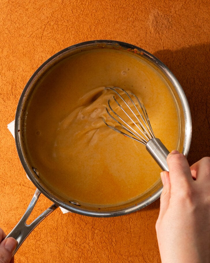 A person uses a whisk to stir a creamy, orange-colored sauce in a stainless steel saucepan placed on an orange surface.